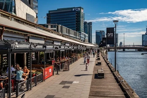 A series of restaurant terraces lined up along the stone quay of a river in Melbourne on a sunny afternoon.